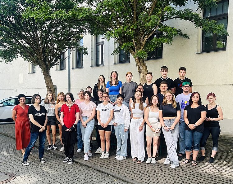 Das neue Schuljahr beginnt Bild mit 23 Schülern und 2 Lehrerinnen, die bei sommerlichem Wetter draußen auf dem Gehweg unter zwei Bäumen für das Klassenfoto posieren.