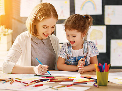 Eine lächelnde junge Frau sitzt an einem Tisch und zeichnet zusammen mit einem kleinen Mädchen, das ebenfalls lächelt. Im Vordergrund sind bunte Stifte und Papier zu sehen, im Hintergrund hängen Kinderzeichnungen an einer Tafel.