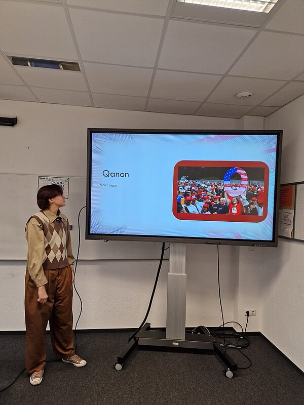 Ein Schüler steht vor einer Folie mit dem Titel „Qanon“. Auf dem Bildschirm ist ein Bild von Menschenmengen mit einer großen Q-Symbolik zu sehen – ein Hinweis auf die bekannte Verschwörungstheorie.