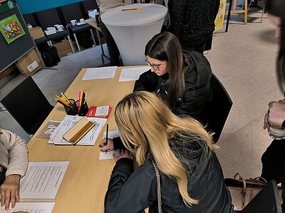 Zwei Personen sitzen an einem Tisch und sind in Dokumente vertieft. Eine Person schreibt, während die andere liest. Auf dem Tisch liegen verschiedene Papiere und Schreibutensilien.