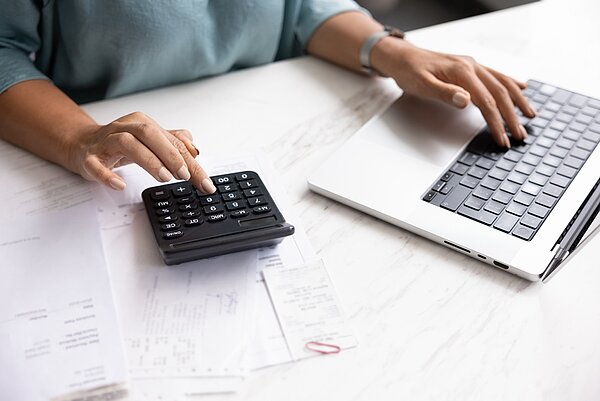 Man sieht die Arme und Hände einer Person, die an einem Schreibtisch sitzt und arbeitet mit einem Laptop sowie einem Taschenrechner, während verschiedene Dokumente und Papiere auf dem Tisch liegen.