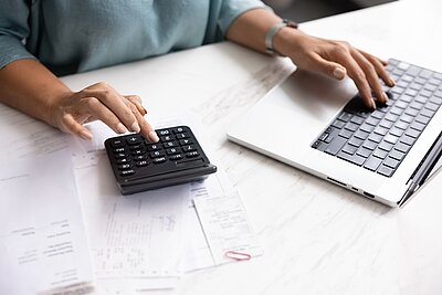 Man sieht die Arme und Hände einer Person, die an einem Schreibtisch sitzt und arbeitet mit einem Laptop sowie einem Taschenrechner, während verschiedene Dokumente und Papiere auf dem Tisch liegen.