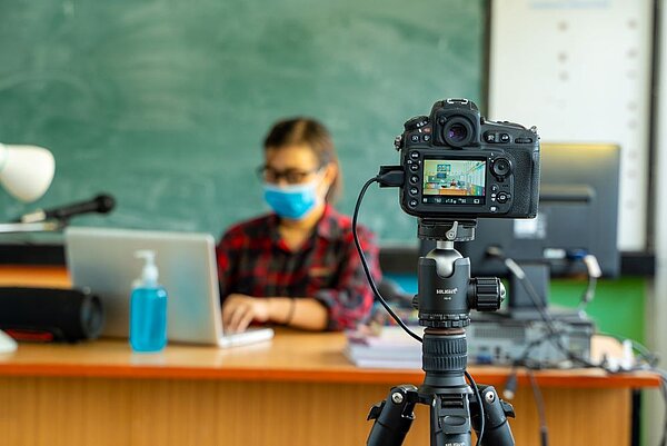 Ton ab! Kamera läuft! Und Action! Eine Lehrerin mit einer blauen Gesichtsmaske sitzt an einem Schreibtisch mit einem Laptop und Notizen, während eine Kamera auf einem Stativ im Vordergrund auf ihn gerichtet ist. Im Hintergrund sind eine grüne Tafel und ein Desinfektionsmittelspender zu sehen.