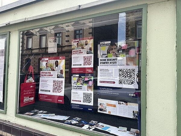 Schaufenster mit mehreren Plakaten und Flyern, die dicht nebeneinander an der Innenseite der Scheibe angebracht sind. Die Plakate zeigen überwiegend weiße Hintergründe mit roten und blauen Akzenten sowie großflächigen QR-Codes. Auf einigen Plakaten sind Porträtfotos von Menschen zu sehen. Die Texte auf den Plakaten informieren über Bildungsangebote und Ausbildungen, unter anderem mit dem Hinweis 'DEINE ZUKUNFT STARTET JETZT!'. Im linken Bereich hängt eine rote Tragetasche mit weißem Aufdruck. Die Spiegelung der gegenüberliegenden Häuserfront ist in der Scheibe sichtbar.