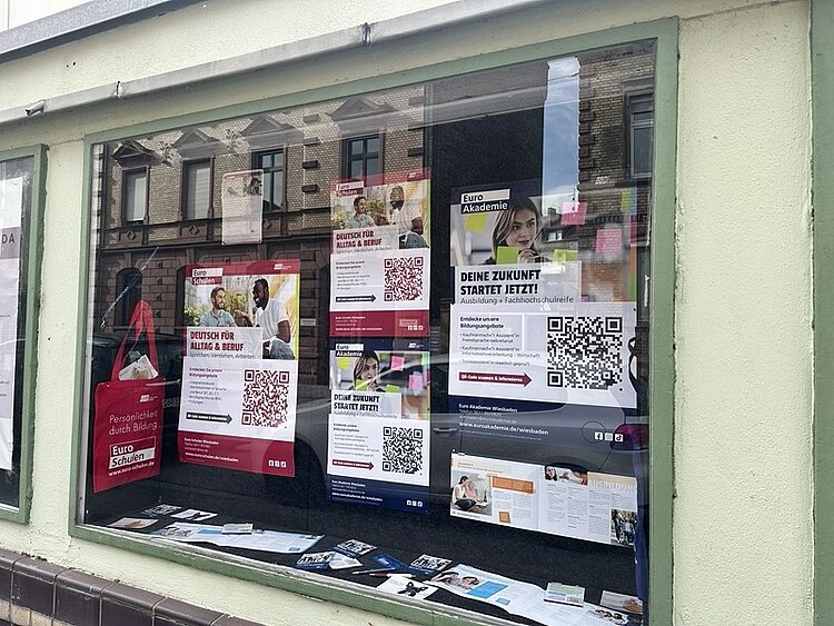Schaufenster mit mehreren Plakaten und Flyern, die dicht nebeneinander an der Innenseite der Scheibe angebracht sind. Die Plakate zeigen überwiegend weiße Hintergründe mit roten und blauen Akzenten sowie großflächigen QR-Codes. Auf einigen Plakaten sind Porträtfotos von Menschen zu sehen. Die Texte auf den Plakaten informieren über Bildungsangebote und Ausbildungen, unter anderem mit dem Hinweis 'DEINE ZUKUNFT STARTET JETZT!'. Im linken Bereich hängt eine rote Tragetasche mit weißem Aufdruck. Die Spiegelung der gegenüberliegenden Häuserfront ist in der Scheibe sichtbar.