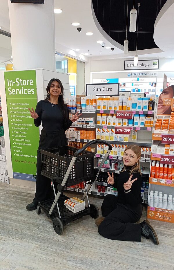 Zwei junge Frauen in einem Drogeriemarkt. Eine steht hinter einem Einkaufswagen, die andere kniet daneben. Beide machen Peace-Zeichen mit den Händen. Im Hintergrund sind Regale mit Sonnencreme und anderen Pflegeprodukten sowie ein grünes Schild mit 'In-Store Services' zu sehen.