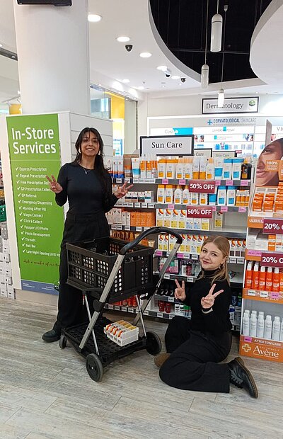 Zwei junge Frauen in einem Drogeriemarkt. Eine steht hinter einem Einkaufswagen, die andere kniet daneben. Beide machen Peace-Zeichen mit den Händen. Im Hintergrund sind Regale mit Sonnencreme und anderen Pflegeprodukten sowie ein grünes Schild mit 'In-Store Services' zu sehen.