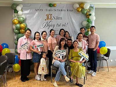Eine Gruppe von Menschen, darunter Erwachsene und ein Kind, posiert lächelnd vor einem Banner mit der Aufschrift „AP09 - Euro Schulen Jena, 10 Năm, Ngày trở về!, Hẹn Uớc Thân”. Im Hintergrund sind Luftballons in Grün, Gold und Weiß sowie ein Raum mit Stühlen zu sehen.