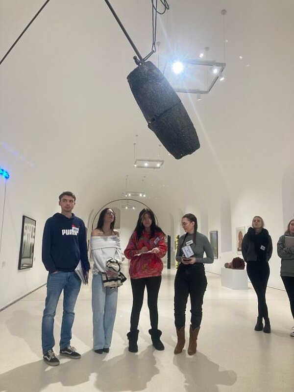 Eine Gruppe von Personen steht in einer Kunstgalerie und betrachtet ein großes, von der Decke hängendes Kunstwerk. Die Galerie hat weiße Wände und ist gut beleuchtet, mit weiteren Kunstwerken an den Wänden.