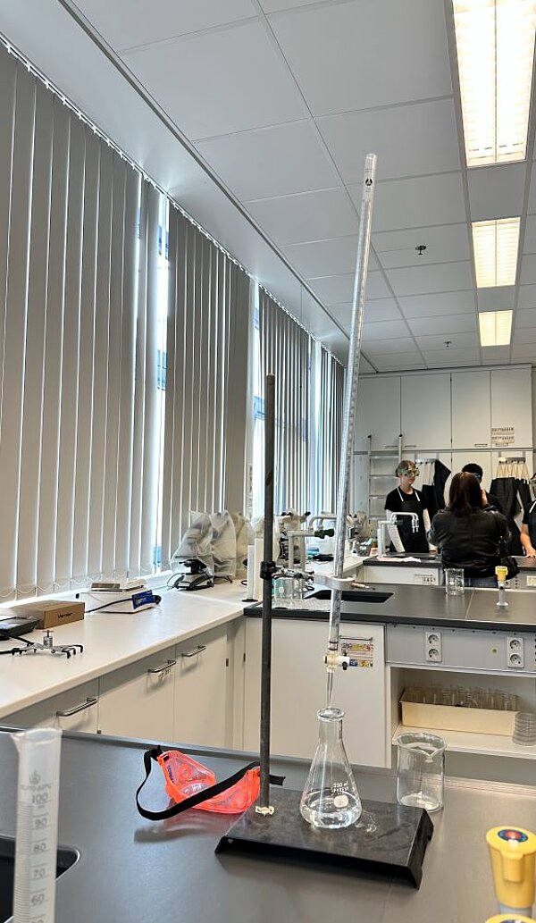 Innenansicht eines Schullabors mit einem Experimentieraufbau, der einen Erlenmeyerkolben und ein Thermometer zeigt. Im Hintergrund arbeiten Schüler*innen an Labortischen mit Mikroskopen und anderen Geräten.