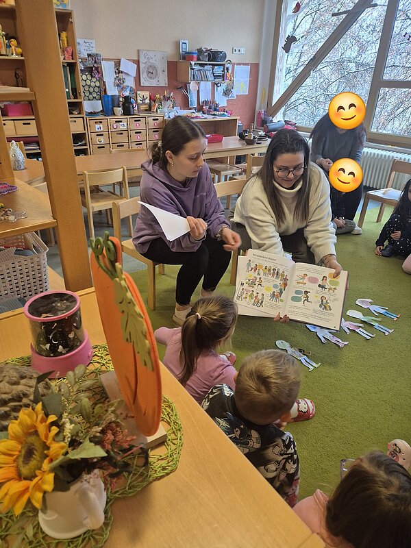 Zwei Vorleserinnen zeigen Kindern in einem Gruppenraum ein geöffnetes Bilderbuch; ausgeschnittene Figuren liegen auf dem Boden.