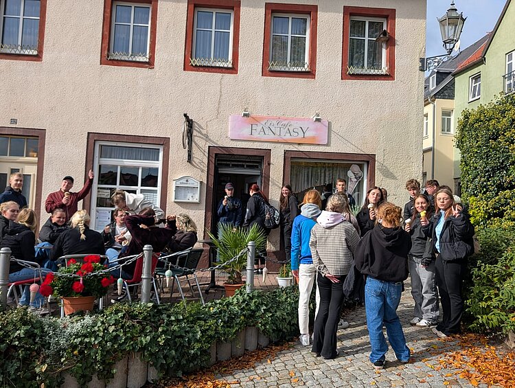 Vor einem Eiscafé mit dem Namen "Fantasy" stehen und sitzen fröhliche junge Menschen mit Eis. Der Platz vor dem Eiscafé ist von Pflanzen gesäumt.