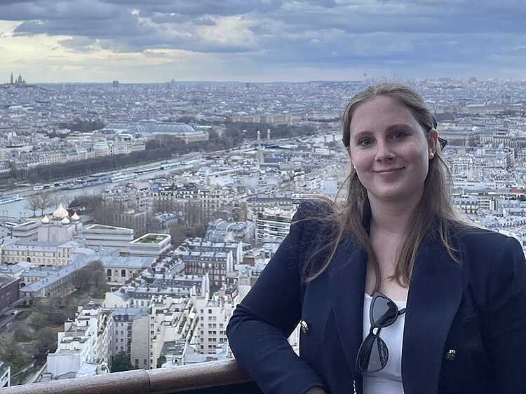 Alumni im Gespräch: Interview mit Caroline Ausmeier Absolventin Caroline Ausmeier posiert für ein Foto, trägt einen marineblauen Blazer und ein weißes T-Shirt, an dem eine schwarze Sonnenbrille angehängt ist.