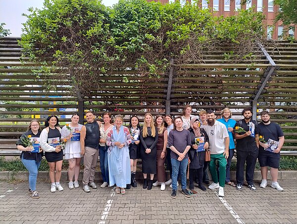 Eine Gruppe lächelnder Menschen steht auf einem Parkplatz vor einer grün bewachsenen Wand aus horizontalen Holzlatten. Einige Menschen halten Blumen und Zeugnisse in den Händen.