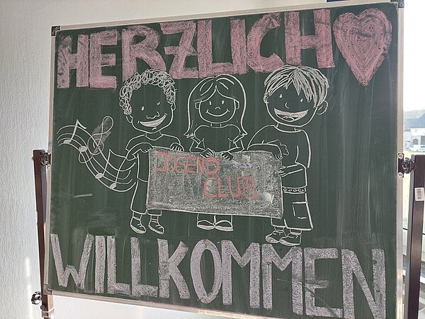 Auf einer großen Tafel an der Wand steht mit Kreide groß Herzlich Willkommen, verziert mit einem Herzen und drei gezeichneten Kinderfiguren