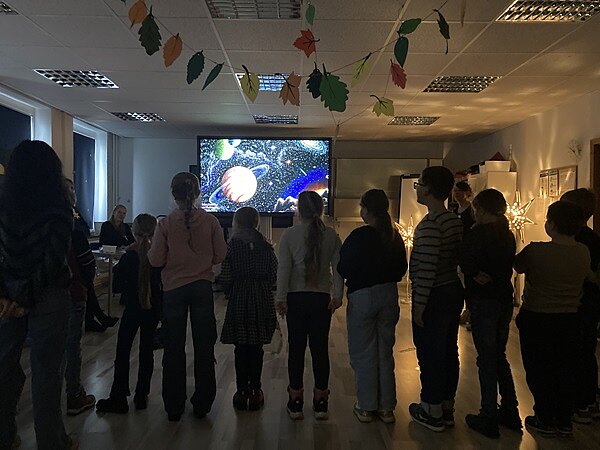 Bundesvorlesetag 2024 Kinder schauen auf den großen Bildschirm, auf dem das Universum und Planeten dargestellt sind.