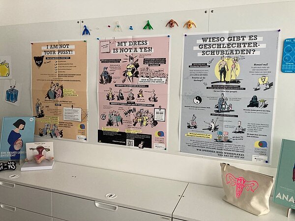 Drei bunte Poster an einer Wand, die über Themen wie Körperautonomie, Kleidung und Geschlechterrollen aufklären.