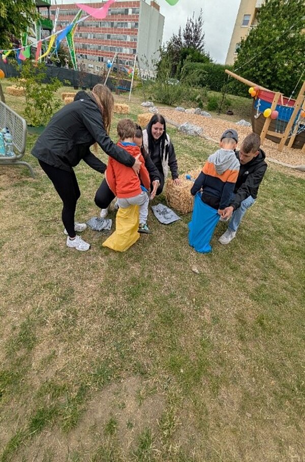 Kinder nehmen auf einer Wiese an einem Sackhüpfen teil und werden dabei von Erwachsenen unterstützt.