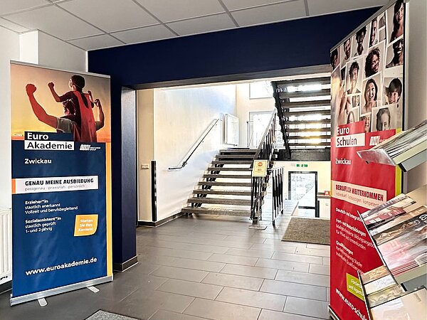 Der Eingangsbereich der Euro Akademie: graue Fliesen, verschiedene Roll-Ups und ein Aufsteller mit Werbematerialien sind zu sehen. Dahinter der blau gestrichene Durchgang, der zum Treppenhaus führt.