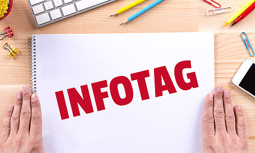 Die Aufschrift "Infotag" in großen roten Lettern steht auf einem Zeichenblock geschrieben, der auf einem Holztisch liegt und neben dem rechts und links die Hände einer Person flach aufliegen. Um den Block verteilt liegen Stifte, Büroklammern und ein Handy und es ist die Ecke einer Tastatur zu sehen.