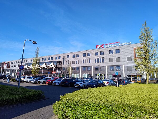 Das Gebäude des MBO College Amstelland in Amsterdam bei sonnigem Wetter. Das moderne Schulgebäude hat eine helle Fassade, und vor dem Eingang sind geparkte Autos sowie ein paar Bäume zu sehen.