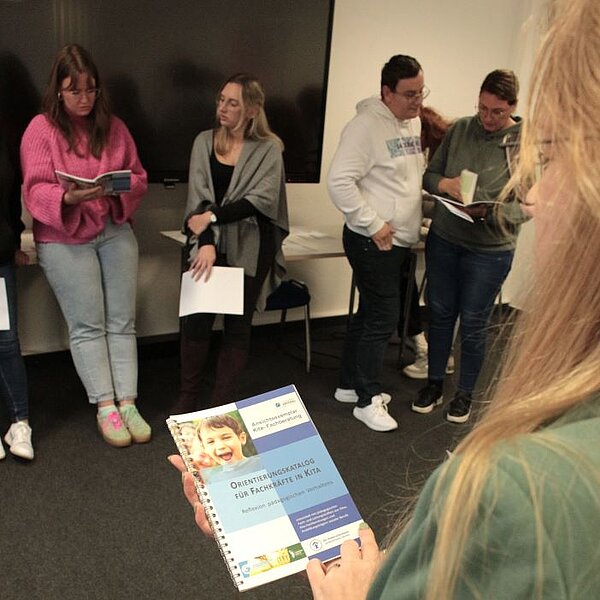 Theorie meets Praxis Eine Person mit langem Haar hält ein Buch mit dem Titel "Orientierungskatalog für Fachkräfte in Kita" in den Händen. Im Hintergrund stehen vier weitere Personen, die ebenfalls in Bücher schauen.