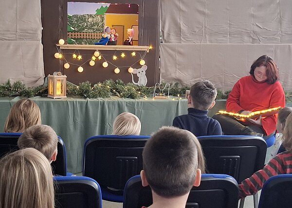 Kinder sitzen auf Stühlen und schauen einem kleinen Puppentheater zu, während eine Frau daneben aus einem beleuchteten Buch vorliest