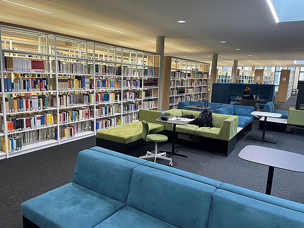 Modern eingerichtete Bibliothek mit blauen und grünen Sofas und Tischen im Vordergrund; im Hintergrund hohe, bunt gefüllte Bücherregale. Einige Personen sitzen lesend bzw. arbeitend an den Tischen, große Fenster sorgen für Tageslicht.