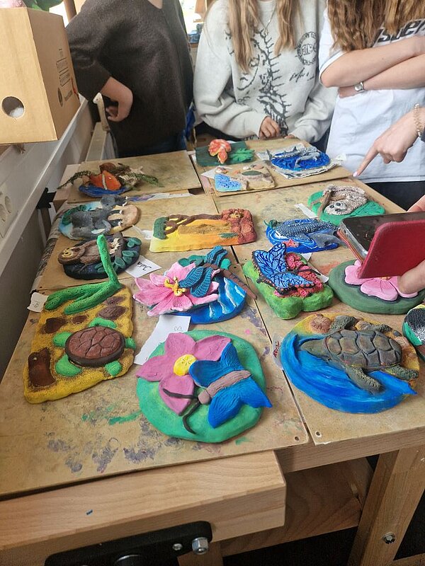 Eine Sammlung von mehreren Tonreliefs auf einer Holzplatte. Die Motive umfassen eine Schildkröte, Blumen mit Schmetterlingen, einen Vogel, eine Eule und vieles mehr. Jedes Relief ist farbenfroh gestaltet und zeigt die kreativen Fähigkeiten der Schüler*innen. Im Hintergrund sind Personen zu erkennen, die um die Platte stehen und auf die Kunstwerke zeigen.