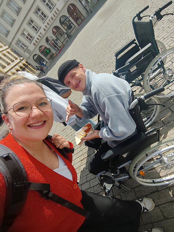 Protesttag zur Gleichstellung von Menschen mit Behinderung: Buttons für Inklusion Das Selfie, das eine Schülerin gemacht hat, zeigt sie stehend und einen Mitschülerin im Rollstuhl sitzend. Der Junge, der eine schwarze Mütze trägt, isst gerade eine Portion Pommes mit Ketchup. Im Hintergrund ist die Markt Apotheke zu sehen.