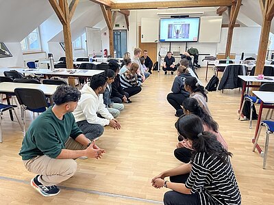 Gruppenaufnahme von Auszubildenden zur Pflegefachkraft an der Euro Akademie Pößneck während eines Gesundheitstages. Die Teilnehmenden stehen in einem Stuhlkreis in einem hellen Schulungsraum mit Holzbalken und großem Bildschirm. Eine Fachperson leitet eine praktische Übung an, die Bewegung, Körperwahrnehmung und Teambuilding fördert. Auf dem Bildschirm im Hintergrund ist eine Präsentation zum Thema Gesundheit zu sehen. Die Szene zeigt einen aktiven, praxisnahen Workshop zu Prävention, digitaler Achtsamkeit und gesundem Arbeiten in der Pflege.