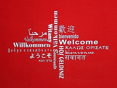 Eine rote Wand mit einem kreuzförmigen Design aus dem Wort 'Willkommen' in mehreren Sprachen, darunter Englisch, Deutsch, Spanisch, Französisch, Arabisch, Chinesisch und weitere.