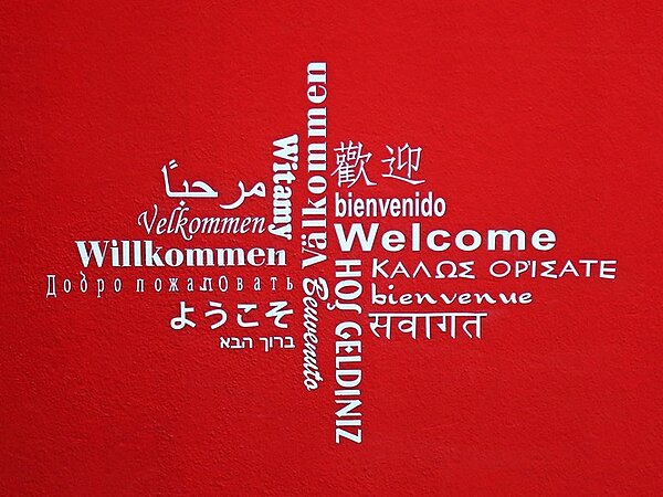 Eine rote Wand mit einem kreuzförmigen Design aus dem Wort 'Willkommen' in mehreren Sprachen, darunter Englisch, Deutsch, Spanisch, Französisch, Arabisch, Chinesisch und weitere.