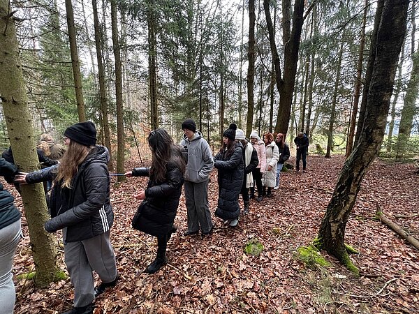 Ein große Gruppe Schüler*innen bewegt sich entlang eines von Baum zu Baum gespannten Seils vorwärts. Die Personen tragen dicke Jacken und teilweise Mützen.