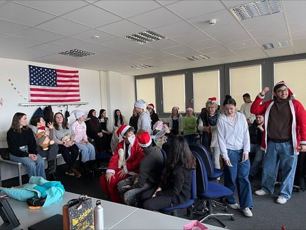 Eine Gruppe von Menschen in einem Klassenzimmer, die sich versammelt haben, um ein Ereignis zu feiern. Einige tragen Weihnachtsmützen und andere festliche Accessoires. Im Hintergrund ist eine amerikanische Flagge zu sehen.