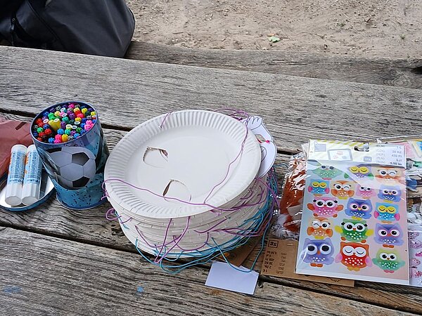 Ein Holztisch im Freien mit Bastelmaterialien: Ein Stapel weißer Pappteller, zwei Klebestifte, ein Paket mit Aufklebern in Form von Eulen und ein blau-weißer Becher mit Fußballmotiv mit bunten Stiften. Im Hintergrund liegt ein schwarzer Rucksack auf einer Holzbank.