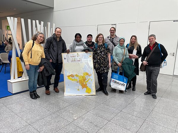 Exkursion zur Messe Eine Gruppe Menschen, die teilweise Jacken in den Händen halten und Taschen dabei haben, befindet sich in einer gefliesten Eingangshalle. Eine Person zeigt eine große Landkarte.
