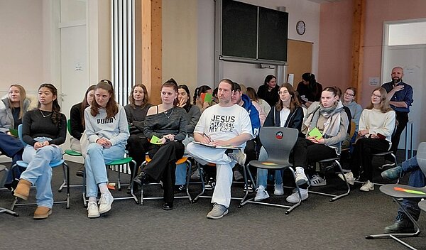 Die Auszubildenden sitzen in einem modernen Seminarraum der Euro Akademie Tauberbischofsheim auf bunten Stühlen in lockerer Anordnung. Im Hintergrund sind eine große Wandtafel, eine Uhr und helle Wände mit Holzsäulen zu sehen. Die Teilnehmenden tragen überwiegend legere Kleidung, darunter Jeans, Pullover und Sneaker. Einige halten bunte Karten in den Händen. Die Szene vermittelt eine offene, kommunikative Atmosphäre im Rahmen eines offiziellen Besuchs zweier Politiker an der Bildungseinrichtung.