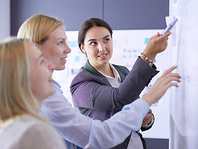 Das Bild zeigt drei Personen, die vor einem Whiteboard diskutieren und schreiben. Es scheint sich um eine professionelle Umgebung zu handeln, möglicherweise ein Büro oder ein Besprechungsraum. Die Personen sind in legerer Geschäftskleidung gekleidet.