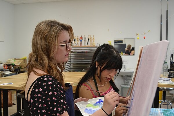 Zwei Besucherinnen des Tages der offenen Tür an der Euro Akademie Würzburg stehen gemeinsam an einer Staffelei, haben Schürzen um und Pinsel in der Hand und malen konzentriert an ihrem Bild. 