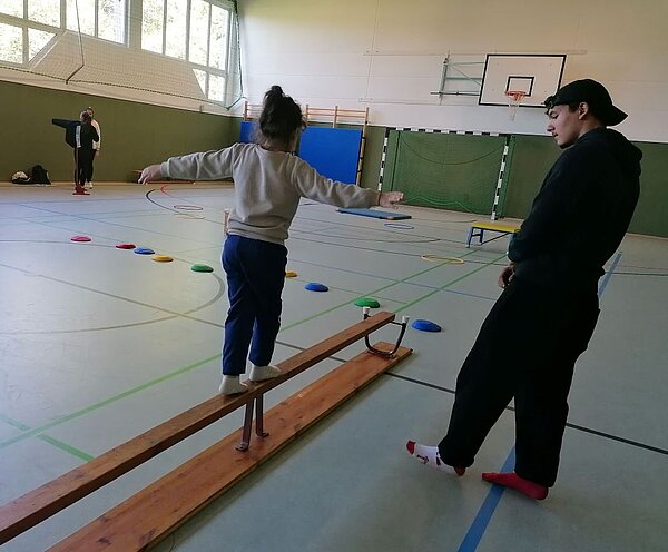 In einer Sporthalle balanciert ein Mädchen mit ausgebreiteten Armen auf einem schmalen Holzbrett auf dem Boden, daneben bunte Übungsscheiben; ein junger Mann mit Basecapsteht hilfsbereit daneben.