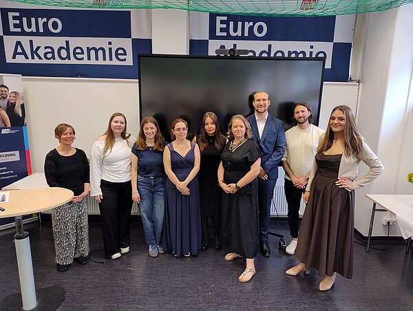 Eine Reihe junger Menschen in festlichen Kleidern steht vor einem großen Bildschirm. An der Wand dahinter ist in dunkelblau zweimal das Logo der Euro Akademie zu sehen.