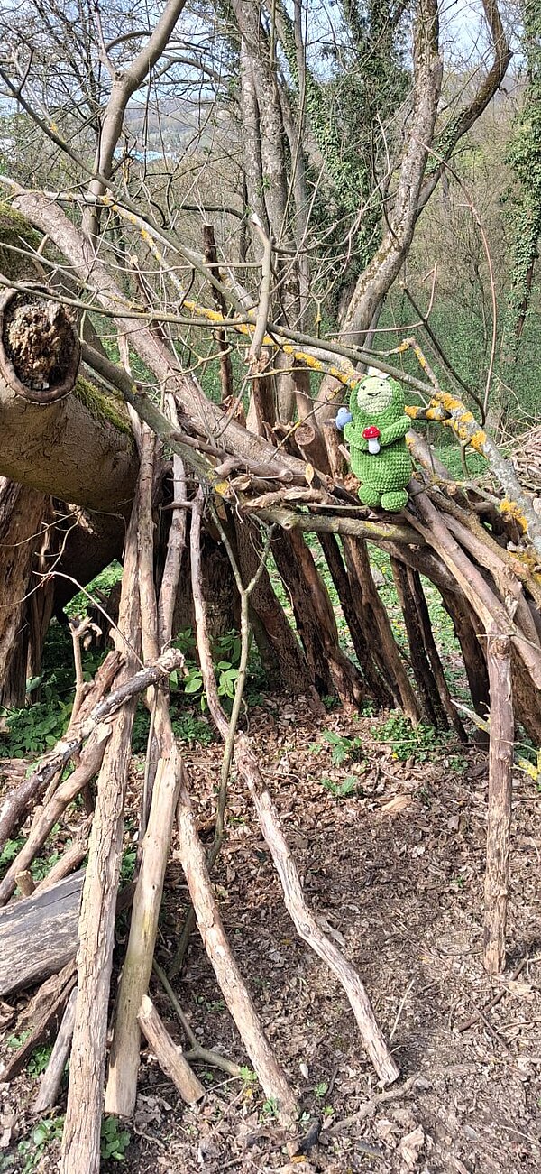 Kleine Konstruktion aus Ästen im Wald, auf der eine grüne gehäkelte Figur mit Pilz sitzt.