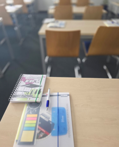 Auf einem Tisch in einem Klassenzimmer liegen Unterrichtsmaterialien, wie einem Schülerkalender, einem durchsichtigen Hefter, der einen Stift, bunte Post-its und einen Schreibblock mit dem Euro Akademie Logo enthält.