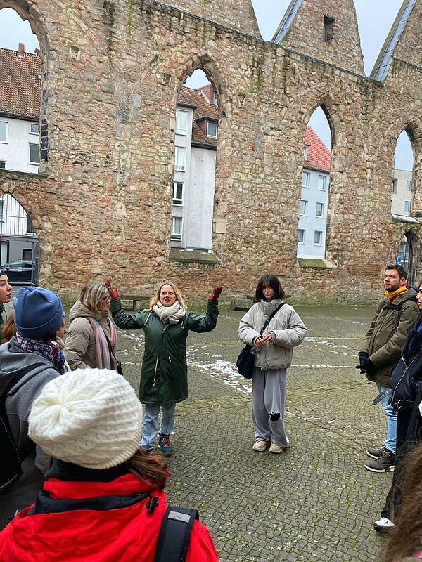Eine Gruppe von Menschen steht vor einer alten Steinmauer mit Bögen und hört einer Frau zu, die mit erhobenen Armen spricht. Die Teilnehmenden sind warm gekleidet, was auf kühles Wetter hinweist. Die Szene deutet auf eine geführte Tour oder einen Vortrag hin, der im Freien vor einem historischen Gebäude stattfindet.