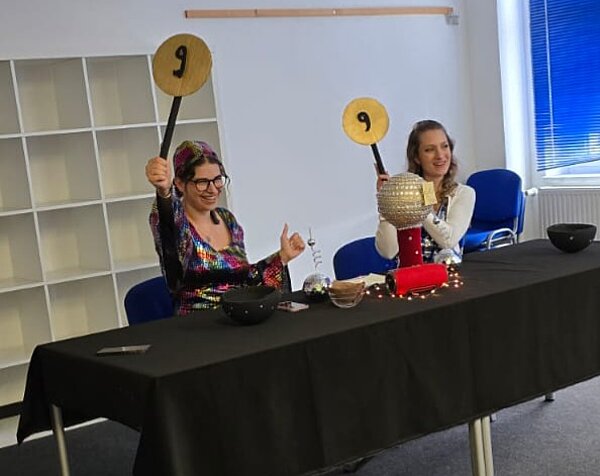 Zwei Personen sitzen an einem Jury-Tisch und halten Bewertungstafeln mit der Zahl 9 hoch.