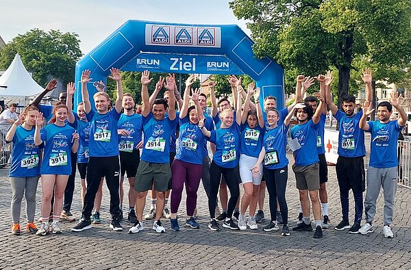 Eine Gruppe junger Erwachsener in sportlicher Kleidung und einheitlichem T-Shirt, worauf ein Aufkleber mit eine Nummer angebracht ist, steht vor dem Zieltor und heben die Arme hoch. Es scheint, als seien sie erfolgreich durch das Ziel gelaufen.