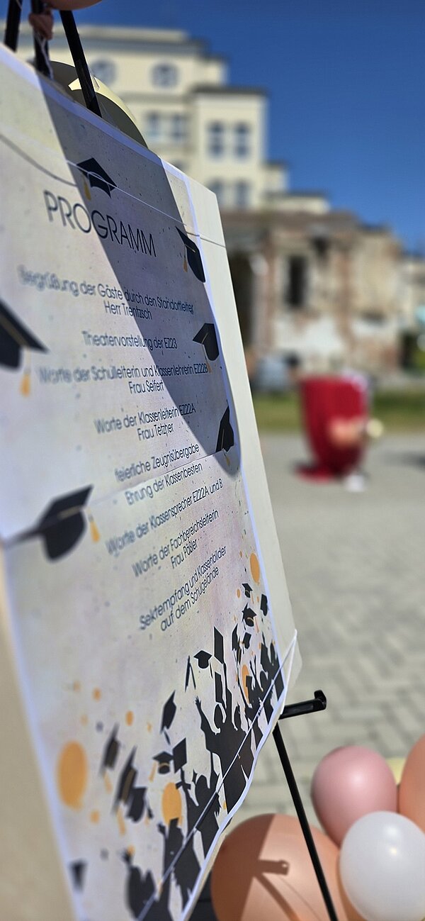 Blick auf das Programm der Abschiedsfeier, das draußen an einer Staffelei hängt, am Boden liegen aufgeblasene Deko-Luftballons.