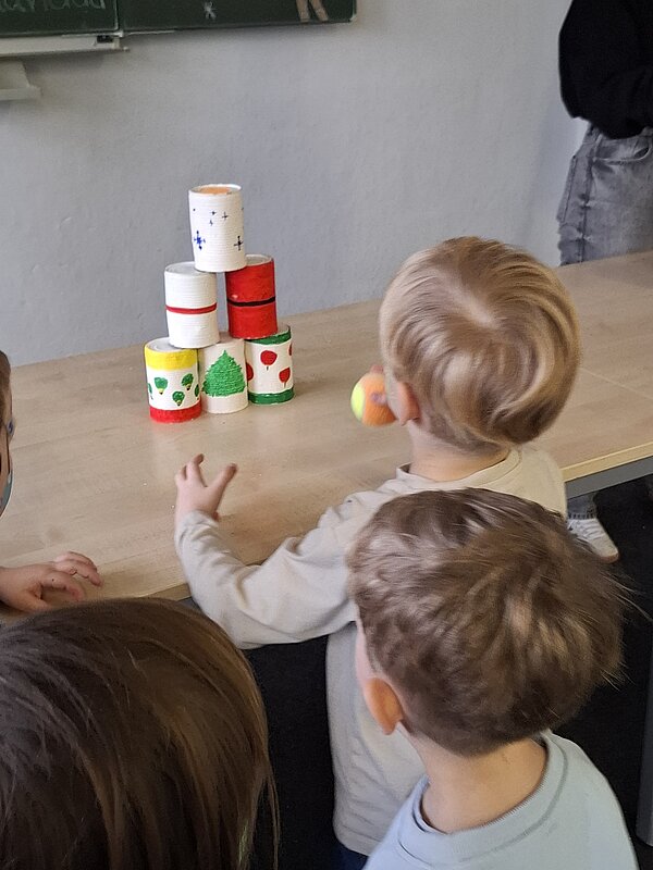 Kinder spielen Dosenwerfen und schauen auf einen Tisch, auf dem bunte, weihnachtlich bemalte Dosen zu einer Pyramide gestapelt sind.