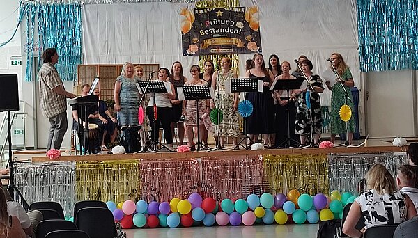Feierliche Zeugnisübergabe an der Euro Akademie Meißen und Euro Akademie Riesa Ein Chor steht auf einer Bühne und singt in Mikrofone. Im Hintergrund hängt ein Banner mit der Aufschrift „Prüfung bestanden!“. Die Bühne ist bunt geschmückt mit Luftballons, Glitzerfolien und Papierblumen. Ein Mann spielt Keyboard und ein anderer trommelt..